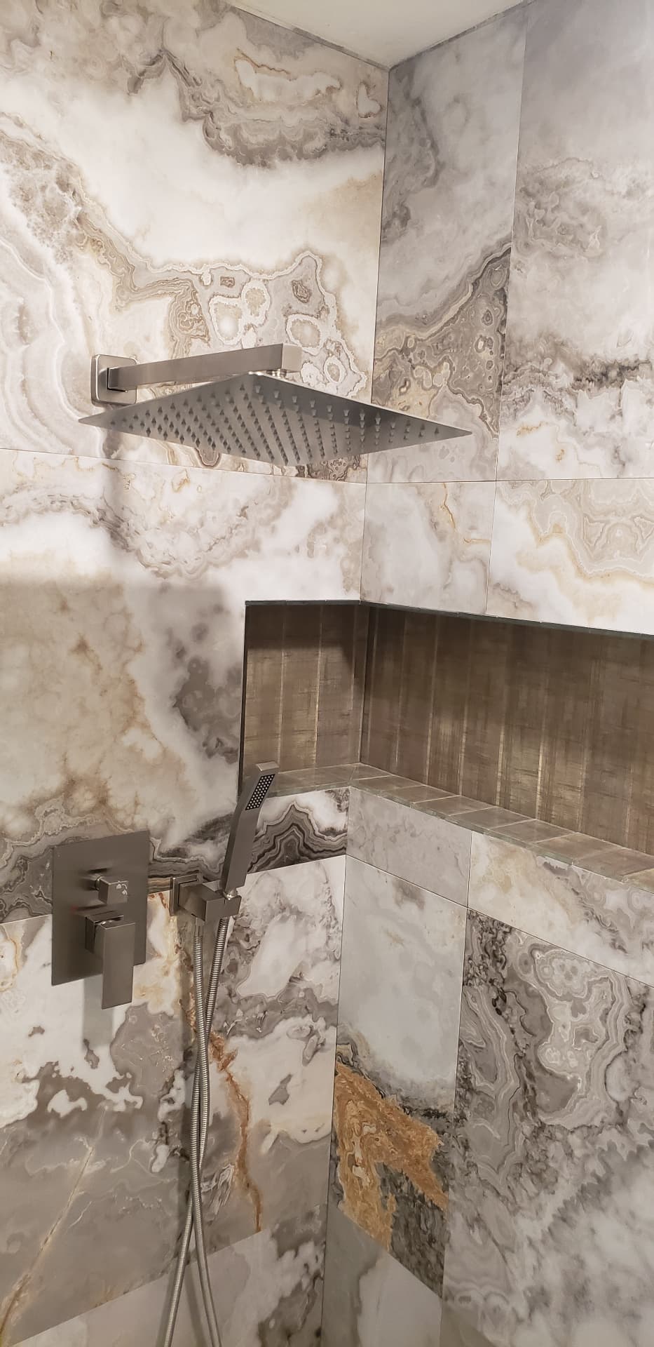Square rain shower head and handheld sprayer against onyx-look marble tile with a recessed niche